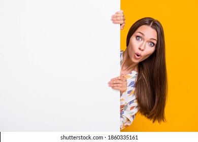 Photo portrait of amazed woman hiding behind white wall with blank space isolated on vivid yellow colored background