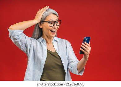 Photo Portrait Of Asian Shocked Amazed Surprised Senior Aged Mature Woman Holding Mobile Phone Seeing Crazy Discounts Wearing Casual Isolated On Red Background.