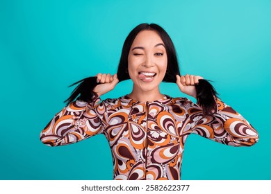 Photo portrait of attractive young woman pulling hair playful wink dressed stylish colorful clothes isolated on cyan color background