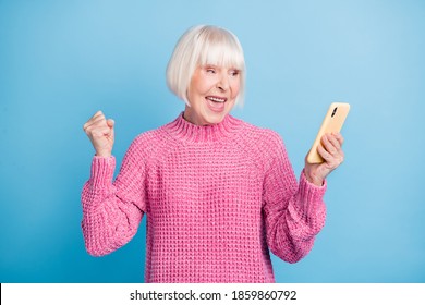 Photo Portrait Of Celebrating Old Lady With Raised Fist Holding Phone In One Hand Isolated On Pastel Blue Colored Background