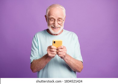 Photo portrait of engaged elderly man holding phone in two hands isolated on vivid purple colored background