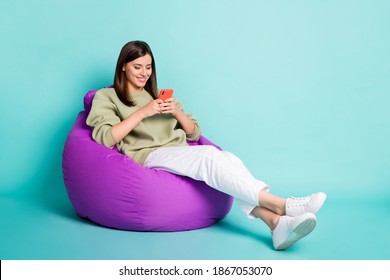 Photo Portrait Full Body View Of Smiling Girl Chatting Holding Phone In Two Hands Sitting In Beanbag Chair Isolated On Vivid Turquoise Colored Background