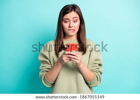 Photo portrait of girl biting lower lip holding phone in two hands isolated on vivid turquoise colored background