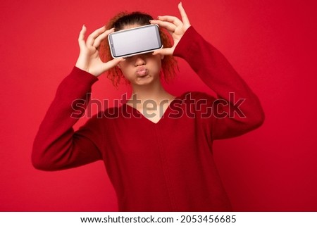 Photo portrait of happy funny young woman wearing dark red sweater isolated over red background holding smartphone and showing mobile phone screen with empty space for mockup having fun