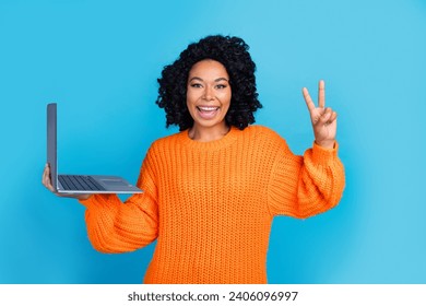 Photo portrait of lovely young lady hold netbook show v-sign wear trendy knitwear orange garment isolated on blue color background