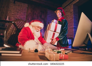 Photo portrait of santa claus writing with quill and ink in scroll and elf carrying three big presents - Powered by Shutterstock