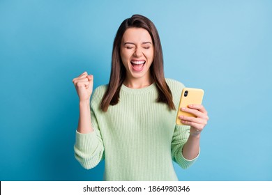 Photo Portrait Of Screaming Girl Celebrating Victory With One Fist In Air Holding Phone In Hand Isolated On Pastel Blue Colored Background