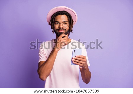 Photo portrait of thinking man in pink headwear holding phone in one hand touching chin isolated on vivid purple colored background