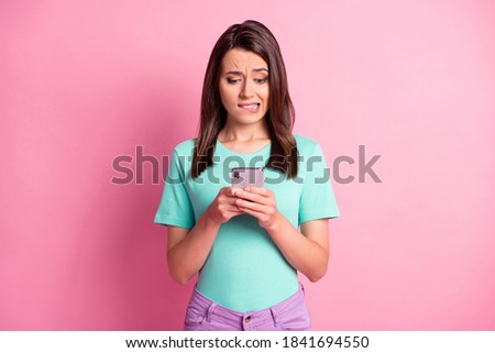 Photo portrait of worried girl holding phone in two hands biting lip isolated on pastel pink colored background