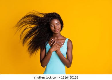 Photo portrait of young curly brunette african american woman holding two hands on chest with closed eyes wearing blue dress isolated on vivid yellow colored background