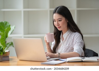 Photo of positive Asian woman holds a cup of coffee imagines plan holiday in office.	