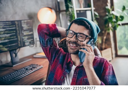 Photo of positive cheerful coder dressed hat glasses smiling talking apple samsung modern gadget indoors workplace workstation loft