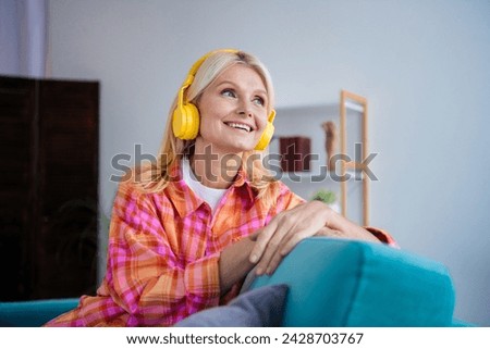 Photo of positive cheerful olf lady sit couch listen wireless headset look dreamy indoors flat