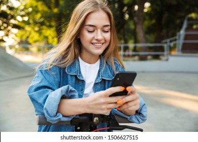 Photo of a positive cheerful teenage girl in park walking on scooter using mobile phone.