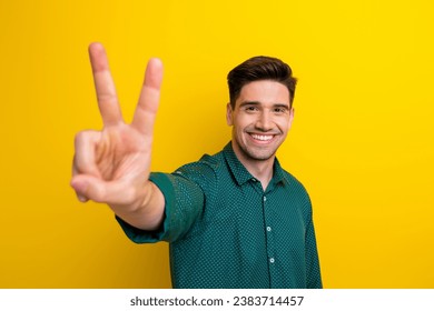 Photo of positive cool guy wear green stylish clothes hand demonstrate v-sign good mood isolated on yellow color background
