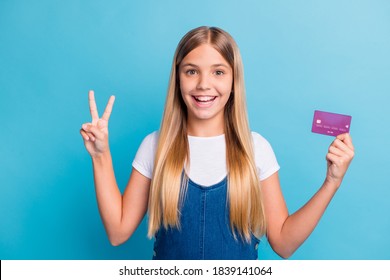 Photo of positive cute blond hair teen girl hold bank card show v-sign wear casual outfit isolated on pastel blue color background