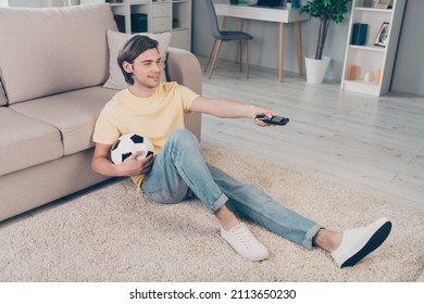 Photo Of Positive Football Fan Guy Sit Carpet Hold Ball Remote Control Switch Channel Wear Yellow T-shirt Home Indoors