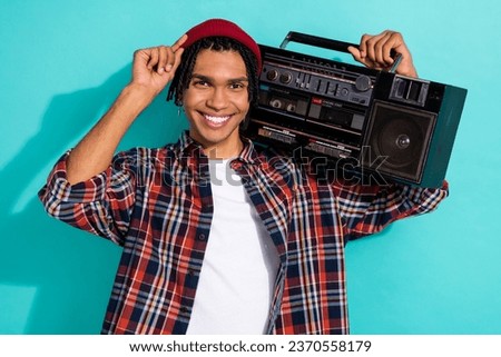 Photo of positive funny optimistic guy with cornrows dressed checkered shirt hold boombox touching hat isolated on teal color background