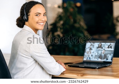 Photo of positive hispanic or brazilian woman with headset, call center operator, turning and smiling at camera, during video conference with multiracial colleagues or clients on laptop screen