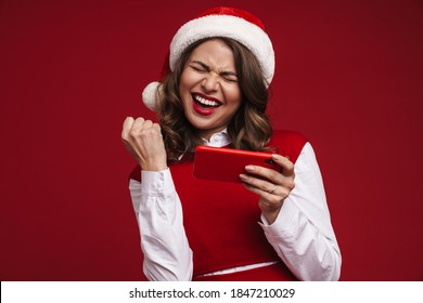 Photo of positive surprised young woman in christmas santa hat using mobile phone and showing winner gesture isolated over red wall background - Powered by Shutterstock