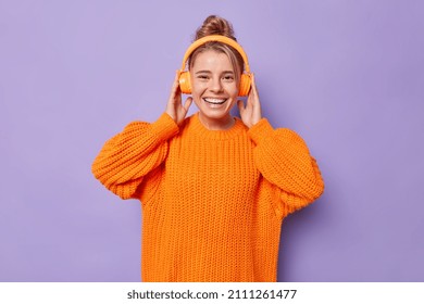 Photo of positive teenage girl has free time for entertainment wears stereo headphones dressed in casual knitted sweater isolated over purple background. People hobby and sincere emotions concept