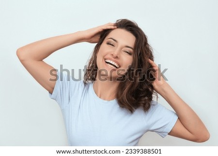 a photo of a positive young happy dark-haired Caucasian woman in a blue T-shirt, who smiling and touching her curly hair with her hands. portrait of a beautiful model. advertisement