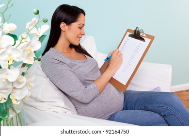 Photo Of A Pregnant Woman At Home Sitting In An Armchair Writing A List Of Possible Boy And Girl Names For Her Baby.