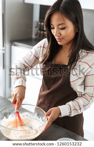 Photo of pretty asian woman 20s smiling and making breakfast in bright kitchen interior