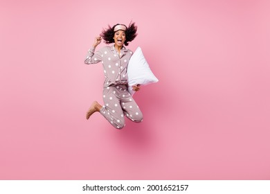 Photo of pretty charming curly dark skin woman dotted nightwear rising fist holding white pillow jumping high isolated pink color background