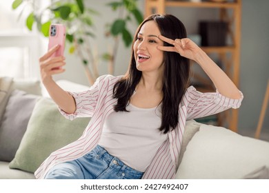Photo of pretty cheerful girl sitting couch hold smart phone make selfie demonstrate v-sign near eye apartment indoors