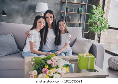 Photo of pretty cheerful lady hug cute daughters sitting on couch toothy smile look camera free time home indoors