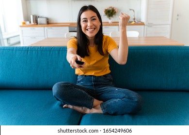 Photo of a pretty cheery positive brunette young woman on sofa indoors at home watch tv make winner gesture.