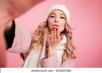 Photo of pretty cute woman standing isolated over pink background wearing warm scarf. Looking camera blowing kisses.