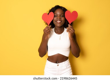 Photo Of Pretty Dark Curly Skin Lady Holding In Hands Little Red Paper Hearts Hiding Flirty Eyes Wear White Casual Clothes Isolated On Yellow Color Background