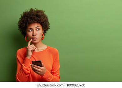 Фотография симпатичной этнической женщины размышляет о том, как ответить на вопрос, глубоко думает о чем-то, пользуется современным мобильным телефоном, пытается составить хорошее сообщение, держит указательный палец возле губ, стоит в помещении