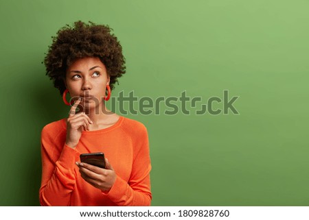 Photo of pretty ethnic woman ponders on how to answer question, thinks deeply about something, uses modern mobile phone, tries to made up good message, keeps index finger near lips, stands indoor