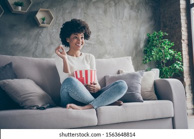 Photo of pretty funny dark skin wavy lady homey mood eating popcorn watching favorite humor television show sitting cozy couch casual sweater jeans outfit flat indoors