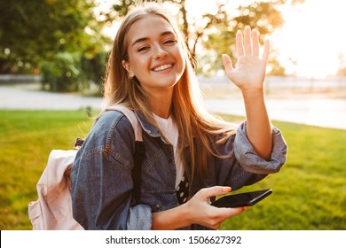 Photo Of A Pretty Happy Young Teenage Girl Walking Outside In Nature Green Park Waving To You Using Mobile Phone.