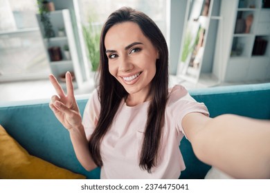 Photo of pretty person sit couch toothy smile make selfie hand fingers demonstrate v-sign free time house indoors