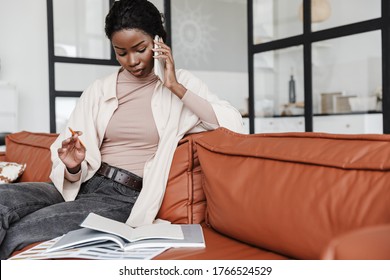 Photo Of Pretty Serious Young African Woman Sitting On Sofa Indoors At Home While Talking By Mobile Phone Near Documents