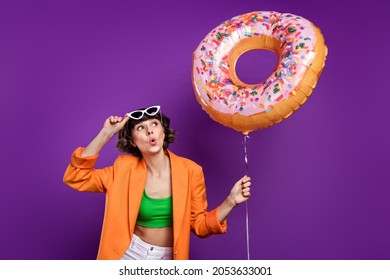 Photo of pretty shocked young lady wear orange jacket arm dark glasses looking holding donut balloon isolated violet color background