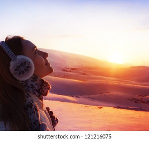 Photo Of Pretty Woman Walking In Snowy Mountains, Side View Of Cute Girl Looking Up, Closeup Portrait Of Female Wearing Warm Winter Earmuff, Red Sunset, Wintertime Sports, Trekking And Hiking Concept
