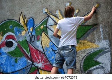 Photo in the process of drawing a graffiti pattern on an old concrete wall. Young long-haired blond guy draws an abstract drawing of different colors. Street art and vandalism concept