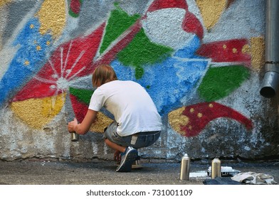 Photo in the process of drawing a graffiti pattern on an old concrete wall. Young long-haired blond guy draws an abstract drawing of different colors. Street art and vandalism concept