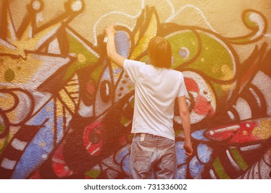Photo in the process of drawing a graffiti pattern on an old concrete wall. Young long-haired blond guy draws an abstract drawing of different colors. Street art and vandalism concept