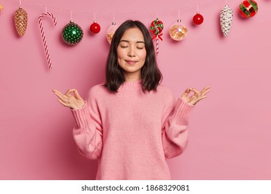 Photo of relaxed brunette woman in casual sweater does meditation gesture with fingers practices yoga on New Year Eve closes eyes has calm expression stands against garland made on christmas toys