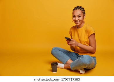 Photo of relaxed dark skinned girl happy to receive message from lover, holds mobile phone, watches funny cute video, sits in lotus pose, drinks takeaway coffee, isolated on yellow background - Shutterstock ID 1707717583