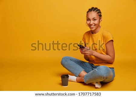 Photo of relaxed dark skinned girl happy to receive message from lover, holds mobile phone, watches funny cute video, sits in lotus pose, drinks takeaway coffee, isolated on yellow background