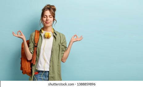 Photo of relaxed student keeps hands in mudra gesture, keeps eyes closed, listens peaceful music, headphones on neck, wears shirt and jeans, has rucksack, isolated on blue background with free space