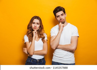 Photo of resented man and woman in casual clothes standing together and touching chin with irritated look isolated over yellow background